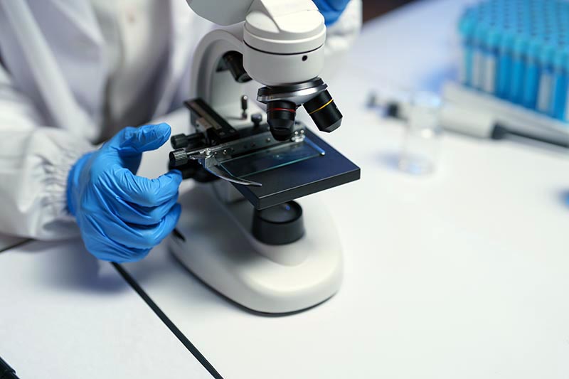 Close-up of gloved hands adjusting a microscope slide on a modern microscope in a medical laboratory, with test tubes and a pipette in the background
